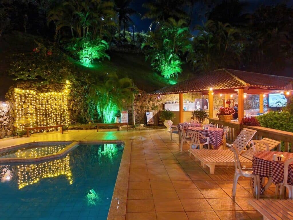 Piscina e bar da Pousada ponta de areia de noite com tudo iluminado, atrás da propriedade há uma montanha e muitas árvores, ao redor da piscina, há cadeiras e mesas