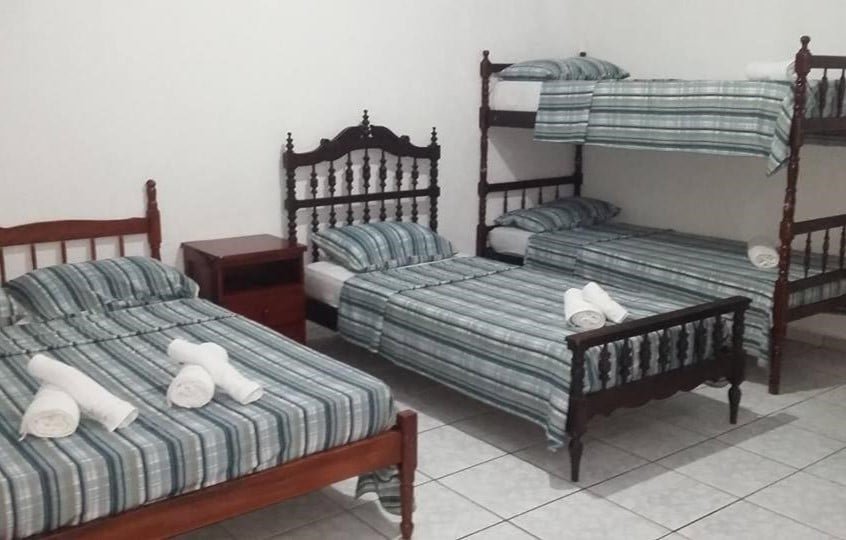 Quarto da Pousada Por do Sol. Uma cama de casal está ao lado esquerdo, uma cama de solteiro está no meio e uma beliche de duas camas está no canto direito. Entre a cama de casal e a de solteiro há uma pequena mesinha com gavetas. 