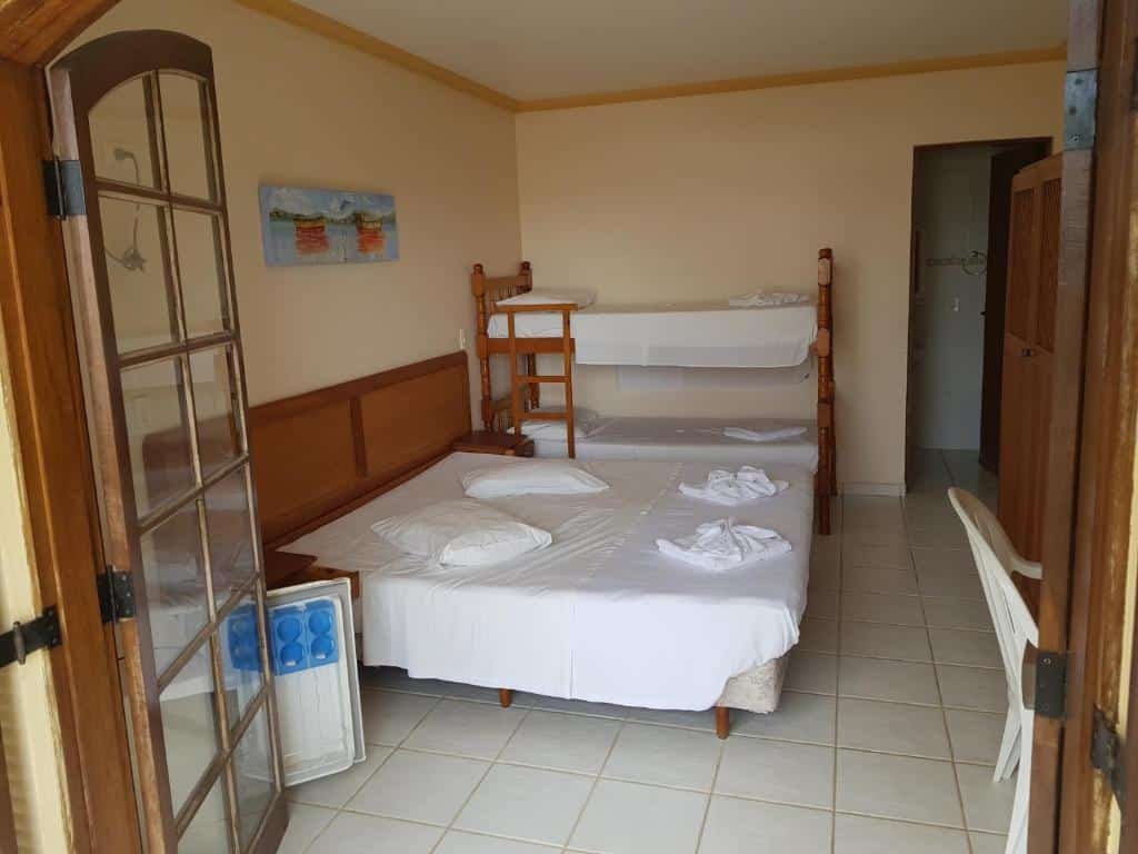 Quarto da Recanto do Morro. Uma beliche de duas camas está encostada na parede do fundo, perto da porta do banheiro. Ao seu lado há uma cama de casal. Em frente as duas há um armário de madeira. Em primeiro plano estão as portas levando para a varanda. 