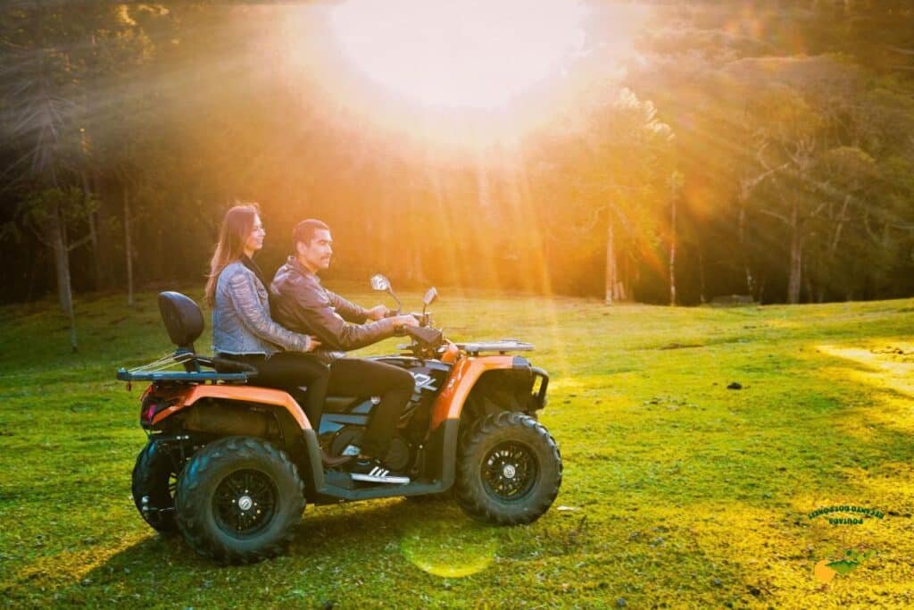 Duas pessoas andam em um quadriciclo laranja em um campo gramado na Pousada Recanto dos Pôneis com a luz do sol brilhando através das árvores ao fundo.