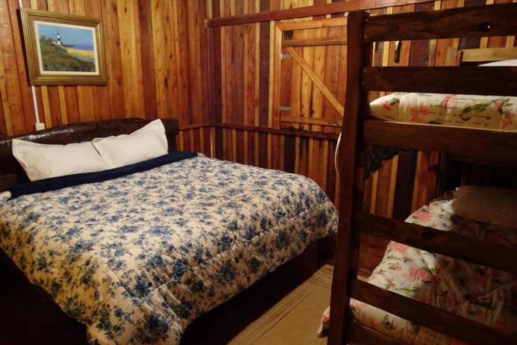 Quarto de madeira com uma cama de casal e beliches na Pousada Recanto dos Pôneis, um dos hotéis fazenda em Santa Catarina. Roupa de cama floral em ambos. Uma pintura de um farol está na parede.