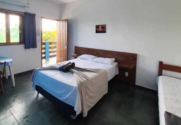 Quarto da Pousada Retiro das Caravelas. Uma cama de casal está ao lado esquerdoa, perto da porta, de uma mesa e da janela. Outra cama está do lado direito. 