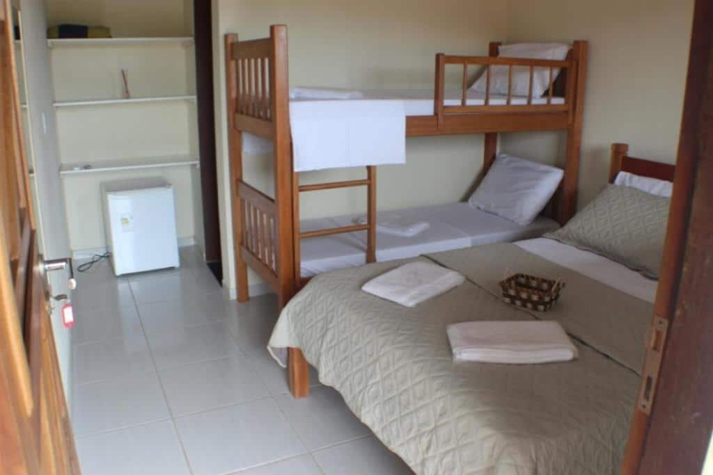Quarto da pousada Pousada Rio Sagi. Uma cama de casal está do lado direito e uma beliche de duas camas está do lado esquerdo. Em frente a beliche existem três prateleiras na parede e um mini frigobar. Essa é mais uma pousada na lista de pousadas em Baía Formosa, RN.