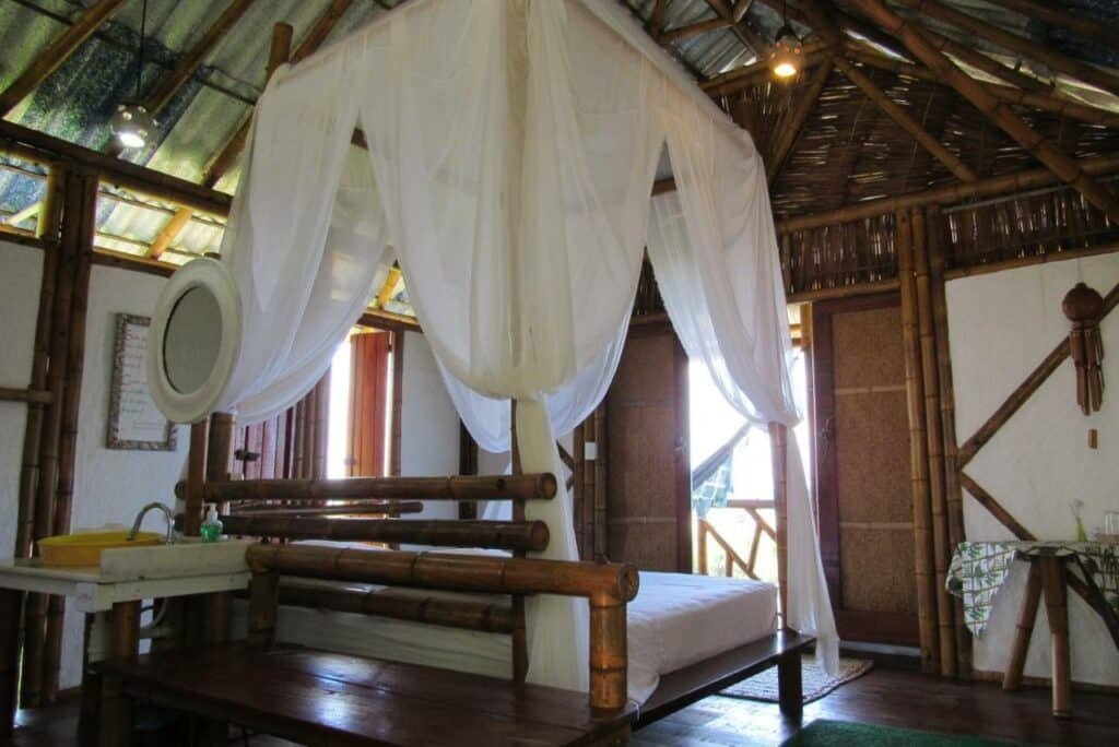 Quarto da Pousada Sabambugi. Uma cama possui um dossel e está no meio do ambiente, virada para a porta aberta. Atrás dela é possíver ver uma mesa e uma pia. Ao lado da porta há outra mesa. Essa é uma das pousadas em Baía Formosa, RN.
