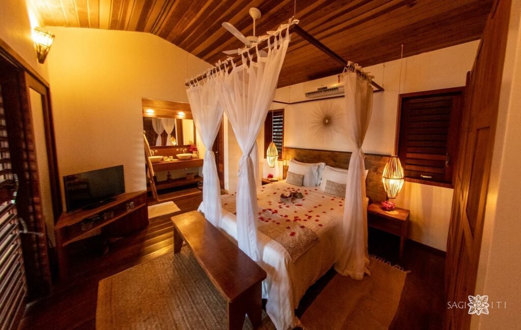 Quarto da Pousada Sagi Iti. Uma cama coberta de pétalas de rosa e com um banco ao seu é está no meio do quarto. Na sua diagonal há uma televisão e ao seu lado esquerdo é possível ver duas pias e um espelho. Aos lados da cama estão também mesinhas e duas luminárias. Está é uma das pousadas em Baia Formosa, RN.