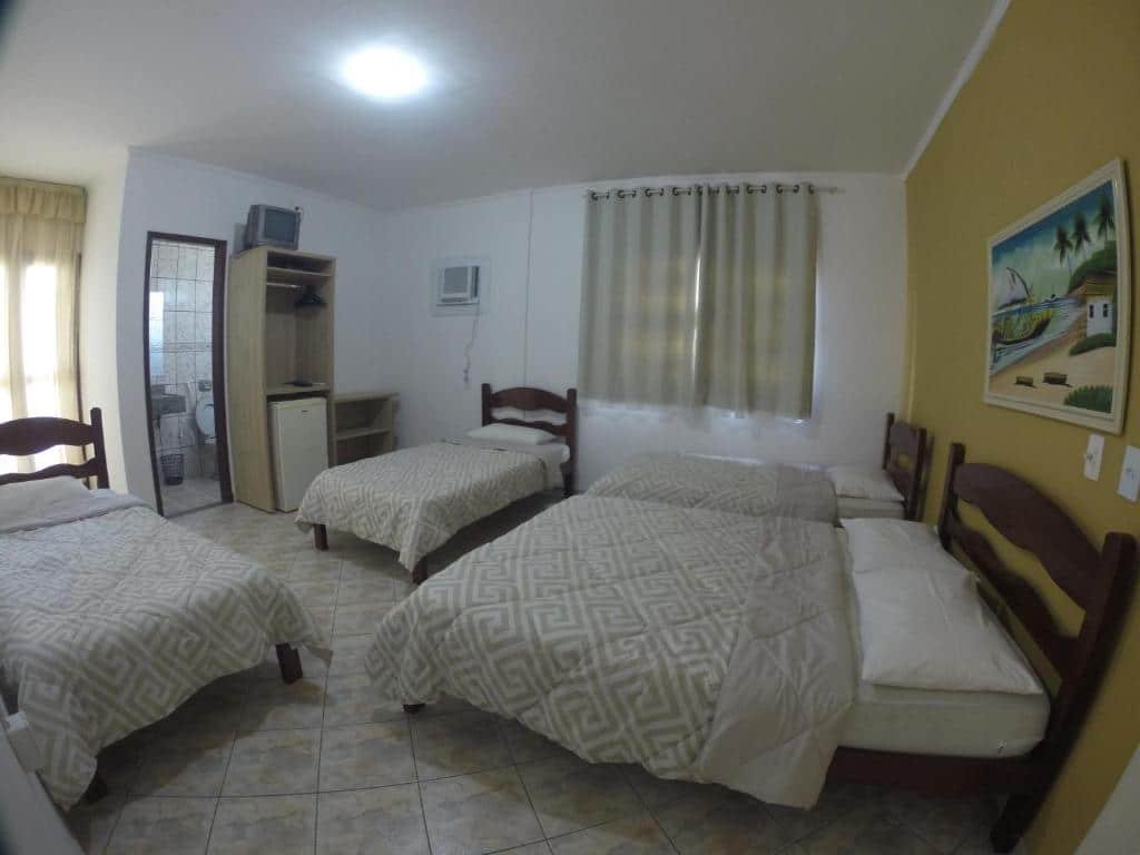 Quarto da Pousada Santa Helena. Uma cama de casal e uma de solteiro no lado direito, de frente uma cama de solteiro e no canto direito outra cama de solteiro. No fundo, no lado direito, um armário, uma televisão antiga e uma porta de banheiro. Foto para ilustrar post de pousadas em Iriri (ES).