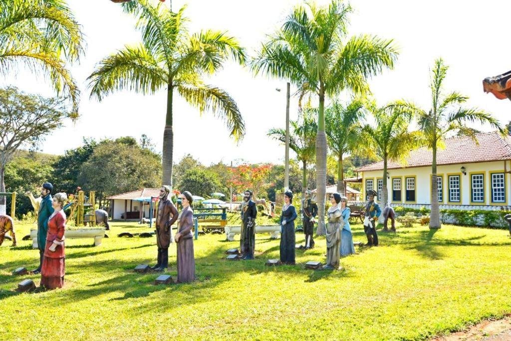 A imagem mostra um jardim gramado com várias estátuas de figuras humanas em trajes antigos, dispostas em pé, como se estivessem participando de uma cerimônia ou encontro. As estátuas estão posicionadas sob palmeiras, e ao fundo há uma construção de estilo colonial, com janelas brancas e molduras verdes. O cenário é ensolarado, com vegetação ao redor, criando uma atmosfera tranquila e histórica. O ambiente parece ser um espaço de exposição ao ar livre, cercado por árvores e outras áreas de lazer ao fundo. Essa é a Pousada temática Estrada Real, um dos hotéis-fazenda em Minas Gerais.