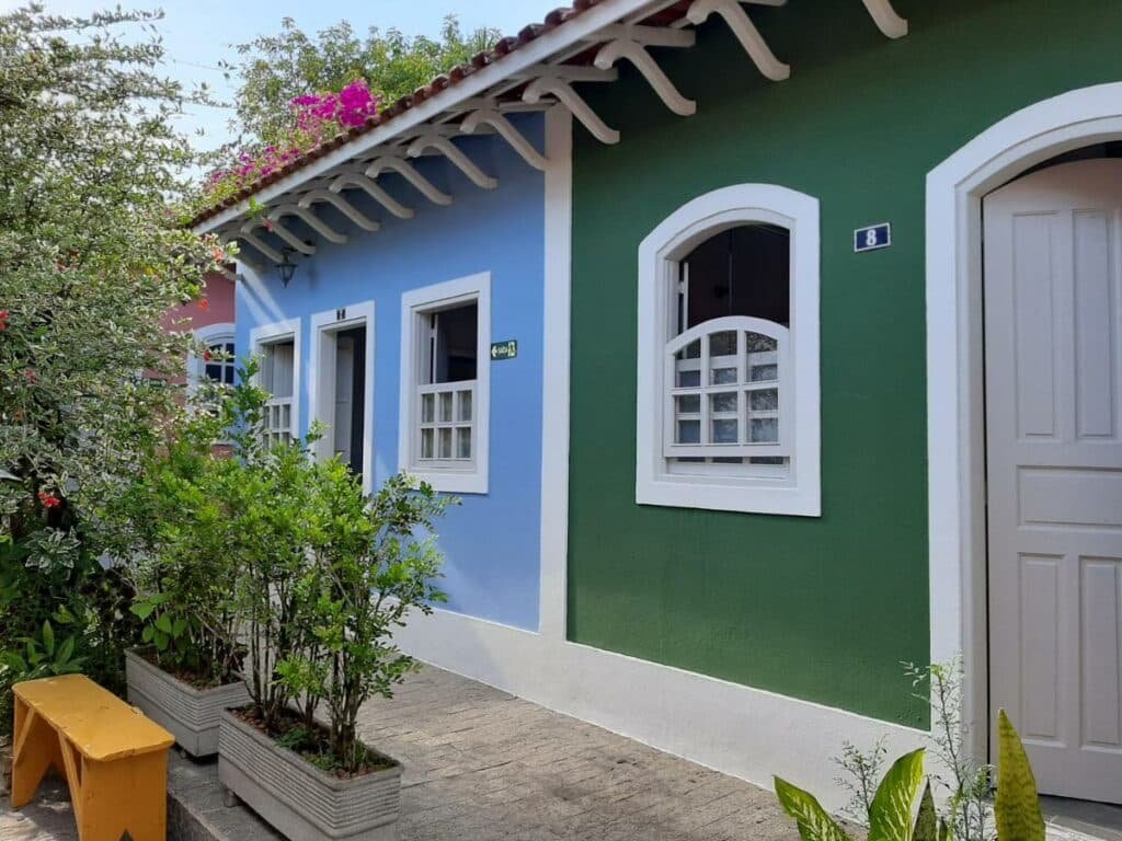 Lado de fora dos quartos da acomodação. Eles possuem portas estreitas e janelas de tamanho médio. Em frente existem várias plantas e flores.