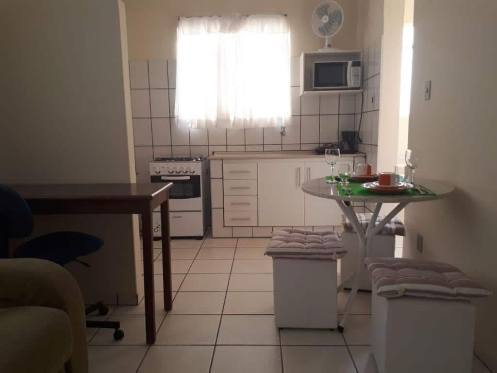 área interna da Pousada Vista da Serra em Ouro Branco, perto de Congonhas, mostrando uma parte da sala de estar e jantar, onde há um sofá, uma mesa redonda com três banquinhos brancos, uma mesa retangular de madeira com uma cadeira preta giratória, e mais atrás - separado por uma parede, é possível enxergar a cozinha equipada com fogão, pia, cafeteira, microondas e ventilador