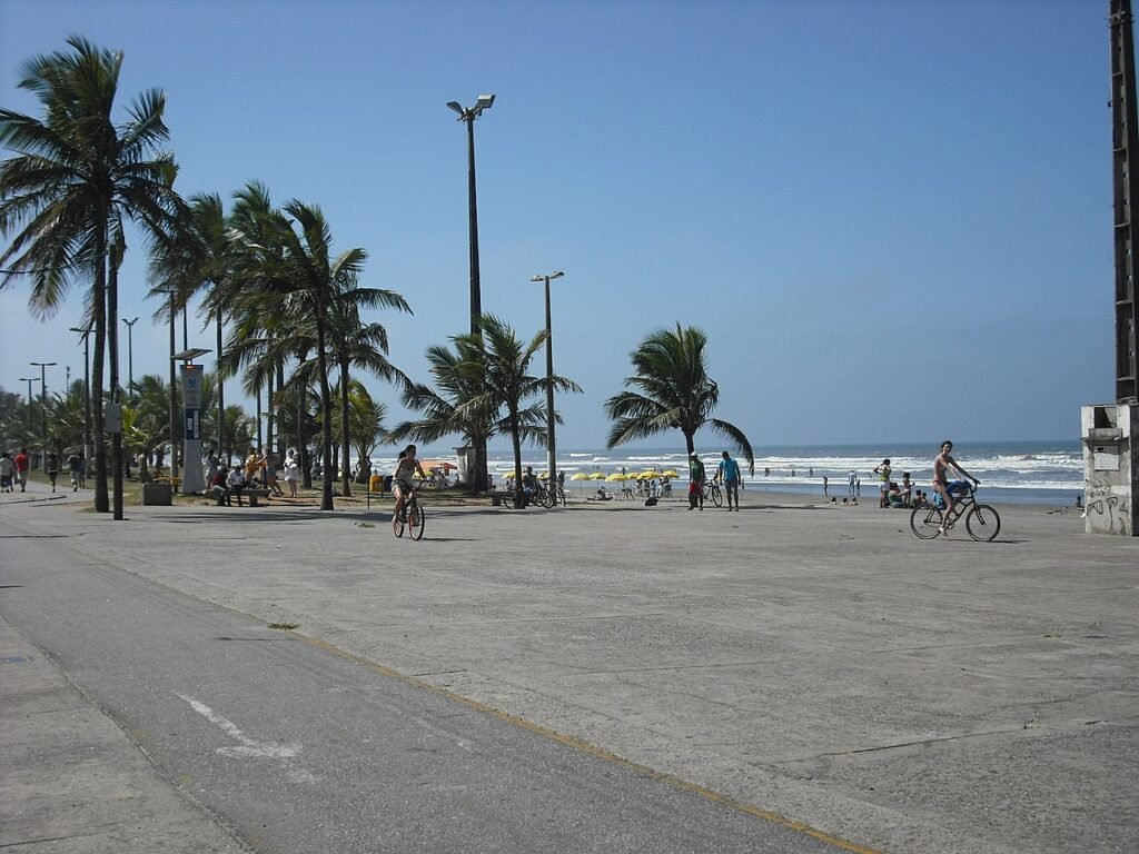 Praia do Centro, em Peruíbe. Um calçadão com ciclovia. Pessoas andando de bicicleta, no lado esquerdo, no fundo, palmeiras e postes de luz. No lado direito o mar com pessoas, guarda-sóis e cadeiras de praia.