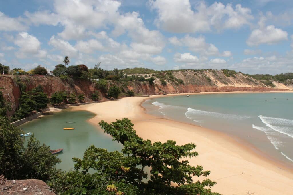 Visão geral da Praia do Porto em Baía Formosa. Ao lado direito está o mar, no meio está a areia e do lado esquerdo uma piscina natural com algumas canoas. Ao fundo é possível ver diversas falésias e árvores.