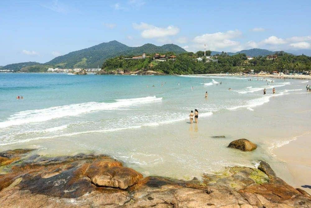 Vista da Praia do Tenório, mar calmo, muitas pessoas, algumas rochas na areia e muita vegetação ao redor