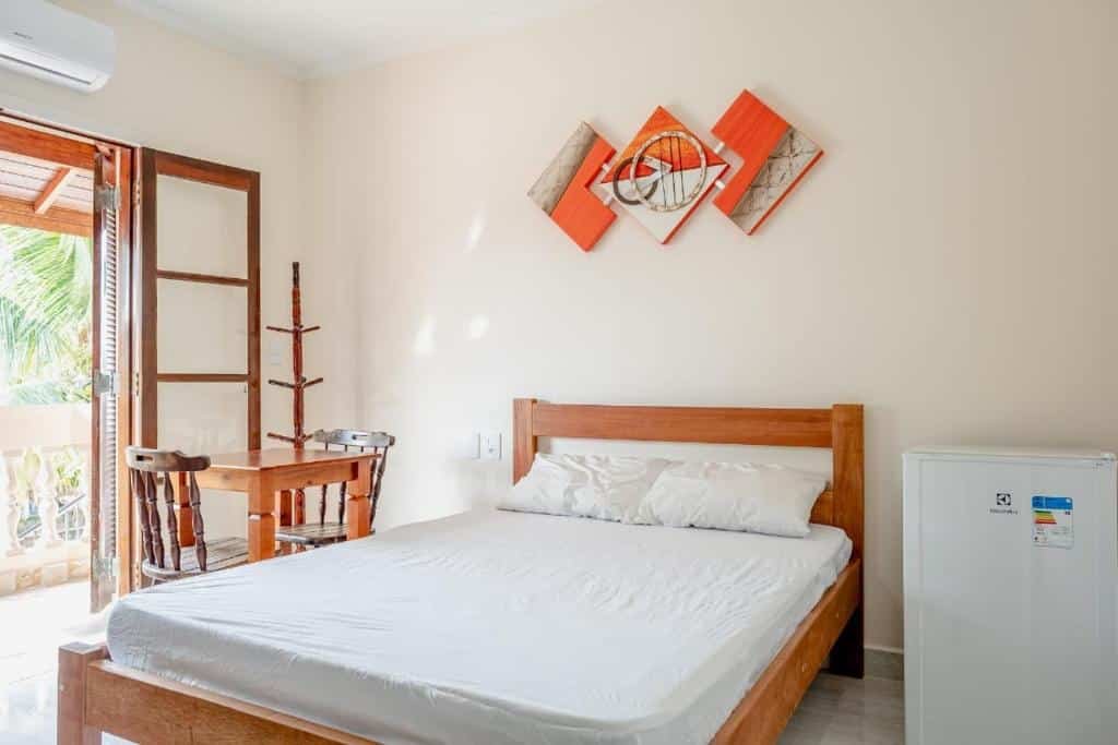 Quarto da Pousada Oásis Café. Um frigobar no canto direito, do lado a cama de casal e do lado esquerdo um cabideiro com uma mesa e duas cadeiras. No lado esquerdo a sacada do quarto.