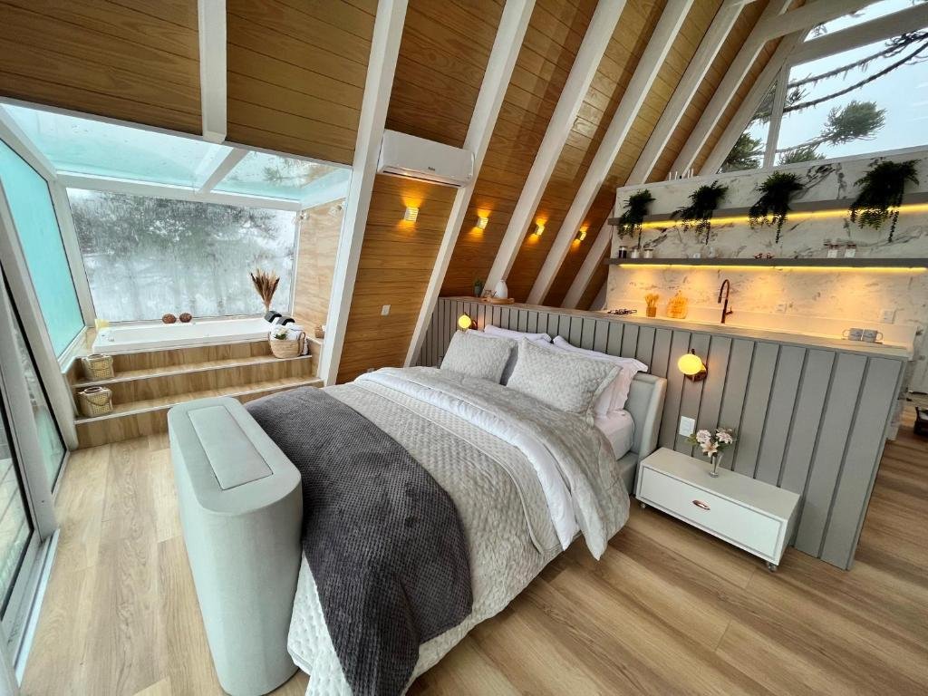 A imagem mostra o interior de um quarto luxuoso no Benta Rosa Chalés de Luxo. O espaço apresenta uma cama de casal grande com roupas de cama brancas e cobertores acolhedores. Atrás da cama, há uma pequena bancada com plantas decorativas e uma pia. O ambiente tem um teto inclinado com estrutura de madeira e grandes janelas de vidro que permitem uma vista para o exterior. Há também uma área elevada com uma banheira de hidromassagem cercada por luz natural, criando um ambiente relaxante e confortável. O estilo é moderno e acolhedor, com tons neutros e acabamentos sofisticados.