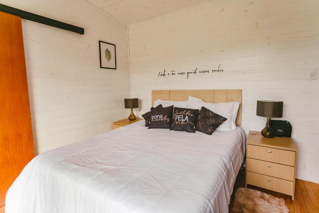 A imagem mostra o interior de um quarto do Chalé Sol Nascente. O espaço apresenta uma cama de casal com roupas de cama brancas e várias almofadas decorativas, duas delas com frases. Ao lado da cama, há dois criados-mudos de madeira, cada um com uma luminária. A parede é de madeira clara, dando um ar acolhedor e rústico ao ambiente. Acima da cabeceira da cama, há uma frase decorativa na parede. O ambiente é simples, aconchegante e organizado.
