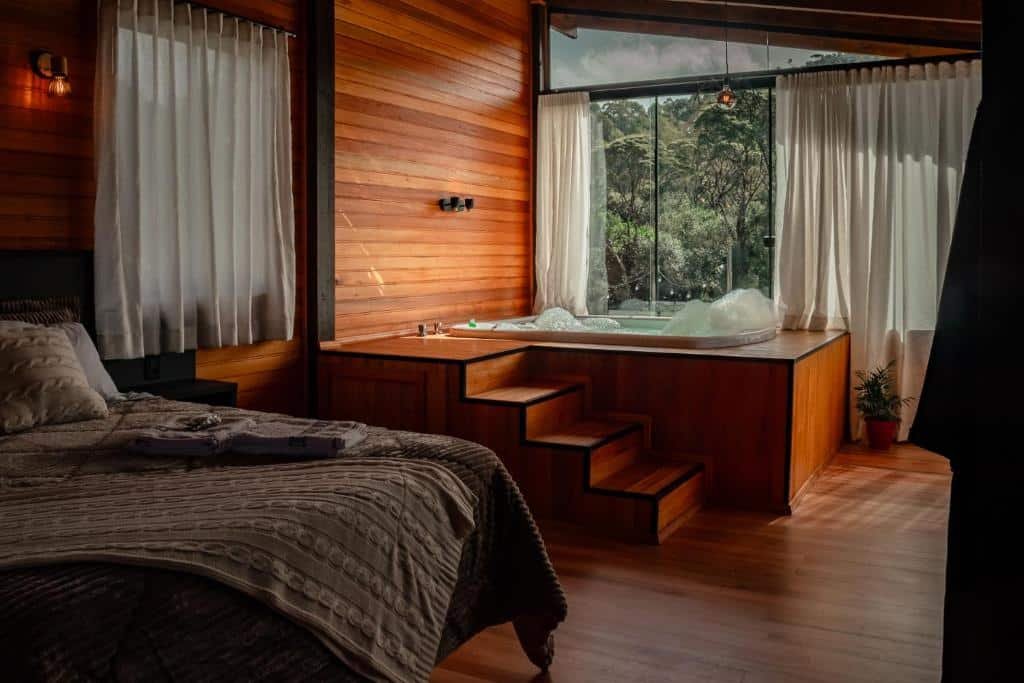 A imagem mostra o interior da Casa de Campo Santa Paulina, um ambiente aconchegante e sofisticado, com paredes de madeira em tom quente. O quarto é equipado com uma cama confortável coberta com uma colcha de tons neutros. Ao lado da cama, há uma banheira de hidromassagem elevada, com degraus de madeira para facilitar o acesso. Grandes janelas de vidro permitem que a luz natural ilumine o ambiente, ao mesmo tempo que oferecem uma vista privilegiada para a vegetação externa. O espaço combina luxo e rusticidade, criando um ambiente perfeito para relaxar e apreciar a paisagem.