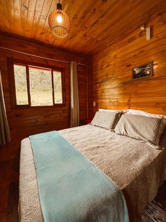 A imagem mostra o quarto da Casa de Campo Santa Paulina aconchegante com paredes e teto totalmente revestidos em madeira de tonalidade quente. A cama de casal está centralizada, com colchas e travesseiros em tons de cinza, e um cobertor azul claro dobrado aos pés da cama, acrescentando um toque de cor. A iluminação do quarto é suave, proporcionada por uma luminária de estilo rústico pendente no teto. Ao lado da cama, há uma janela de madeira com cortinas claras, permitindo a entrada de luz natural e oferecendo vista para a vegetação externa, o que traz uma sensação de proximidade com a natureza. 