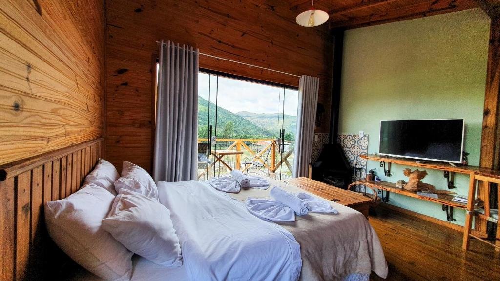 Quarto do Cascata dos Amores, um dos chalés na Serra Catarinense, com cama de casal localizada ao centro, há uma tv em frente, juntamente com prateleiras de madeira, do outro lado, há uma sacada com portas de vidro e mesa com cadeiras de madeira com vista para a natureza.