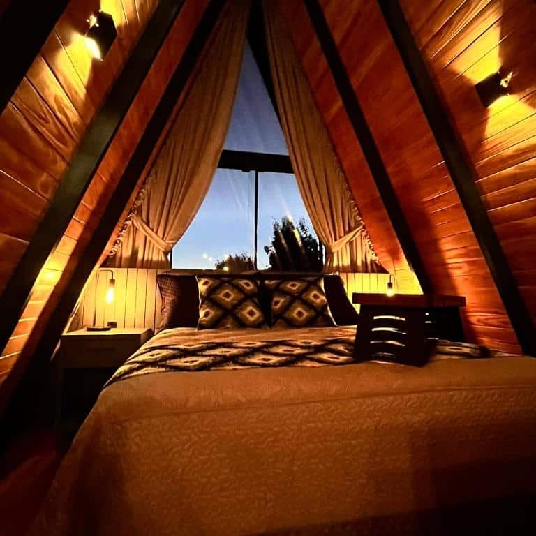 O interior do chalé no Chalé Alqualondë - Chácara Valfenda exibe um ambiente acolhedor e intimista, com iluminação suave que destaca o aconchego das paredes de madeira em formato "A". A cama de casal, centralizada no espaço, está decorada com almofadas de estampa geométrica e uma colcha macia, proporcionando uma sensação de conforto. Ao fundo, uma grande janela triangular com cortinas abertas permite a vista do céu escurecendo ao cair da noite, trazendo a sensação de tranquilidade. O espaço é iluminado por pequenas lâmpadas ao lado da cama, criando um ambiente quente e sereno, ideal para relaxar.