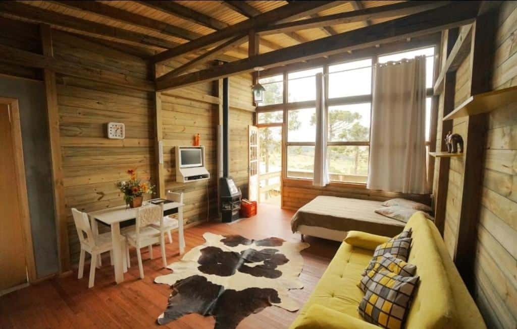 Quarto amplo do Chalés da Serra Catarinense com cama de casal em um canto abaixo de uma parede enorme de vidro com estrutura de madeira. Há uma cortina acima, sofá do outro lado em frente a uma mesinha com cadeira com tv pequena e retrô. No chão, um tapete imitando pele de vaca malhada.