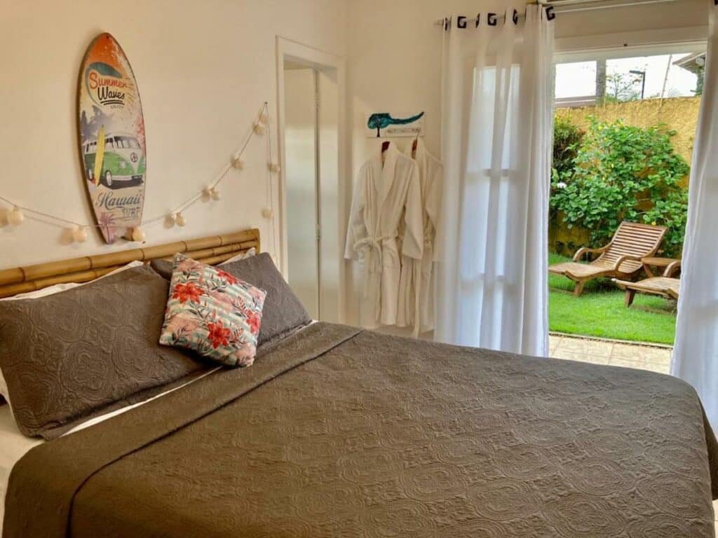 Quarto na MOA Pousada com uma cama de casal, uma varanda dando vista para o gramado e duas cadeiras de madeira, tudo decorado com objetos que remetem a praia