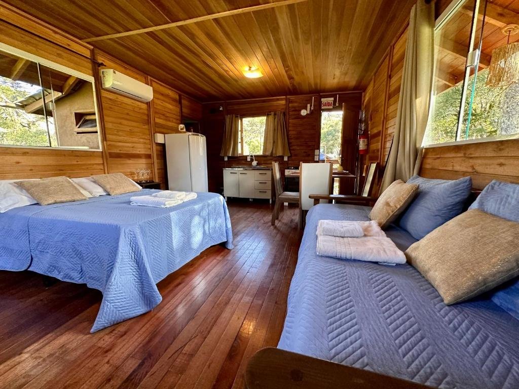 A imagem mostra o interior de um chalé do Namastê - Hospedaria de Altitude. O ambiente é acolhedor, com revestimento de madeira nas paredes e no teto, proporcionando uma sensação rústica e aconchegante. O quarto conta com uma cama de casal, coberta por uma colcha azul, além de um sofá-cama na mesma cor, com almofadas para maior conforto. À esquerda, há uma janela que oferece vista para o exterior, trazendo luz natural ao ambiente. No canto direito, é possível ver uma pequena cozinha equipada com geladeira, pia e mesa para refeições.