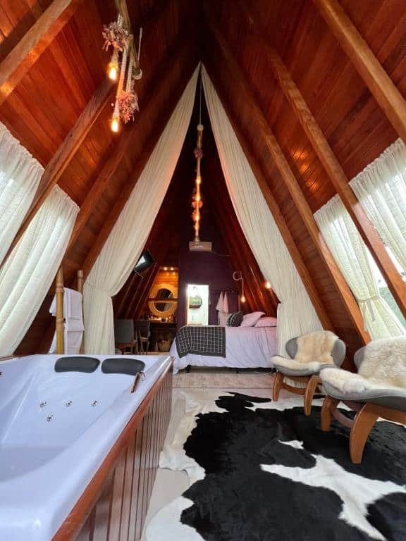 A imagem retrata o interior da Pousada Maçã Araucária, revelando um ambiente sofisticado e rústico ao mesmo tempo. O teto em madeira forma uma espécie de triângulo invertido, com cortinas brancas esvoaçantes penduradas nas laterais, criando uma atmosfera romântica. No centro, uma cama com roupa de cama em tons neutros e uma decoração aconchegante. Em destaque à frente, há uma banheira de hidromassagem em madeira, enquanto no canto direito estão dispostas duas poltronas de design curvo, com assentos de pele, sobre um tapete com estampa de vaca. As luzes pendentes no teto adicionam um toque de calor e charme ao ambiente intimista.