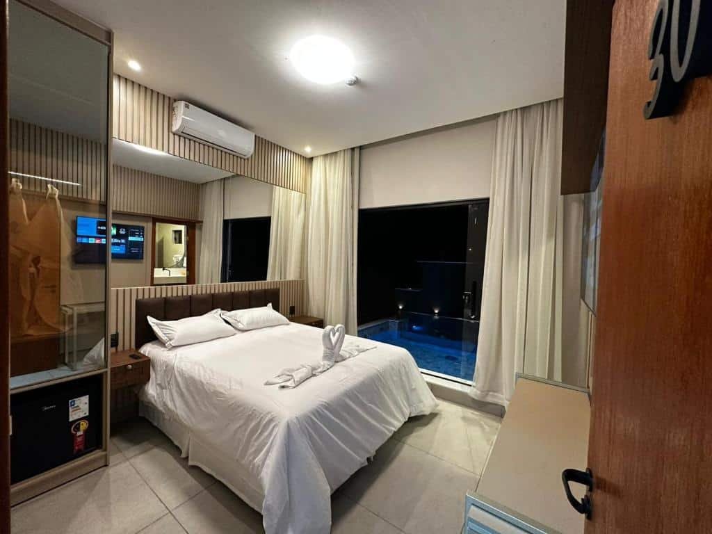 A imagem mostra o interior de um quarto da Pousada Villas em Bonito, Mato Grosso do Sul. O quarto é decorado de maneira moderna e aconchegante, com tons neutros e detalhes em madeira. A cama de casal é coberta com roupas de cama brancas impecáveis e há uma toalha dobrada em forma de cisne sobre a cama.
À esquerda, há um armário com portas de vidro espelhado e um frigobar embutido. Acima do frigobar, há um suporte para malas. Na parede oposta à cama, há uma televisão de tela plana montada, permitindo entretenimento para os hóspedes. O quarto possui ar-condicionado montado na parede para maior conforto.
Uma grande janela com cortinas claras oferece uma vista para a área externa, onde se pode ver uma piscina privativa iluminada à noite, proporcionando um ambiente relaxante e luxuoso.