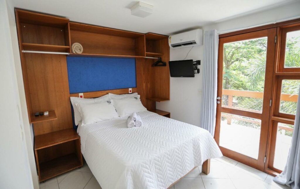 Quarto do Alapage Toque Toque com cama de casal do lado esquerdo da imagem e do lado esquerdo da cama com TV presa na parede. Representa airbnb na praia de Calhetas.