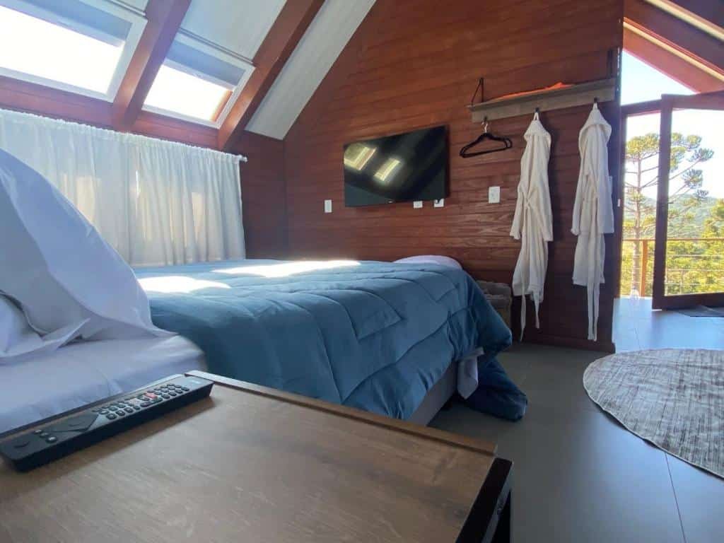 Vista do interior do chalé em Urubici Altitude Máxima com cama de casal e tv em frente, e dois roupões pendurados ao lado, mostrando a arquitetura em estilo alpino. Em primeiro plano, há uma mesa de madeira, combinando com o restante da decoração.
