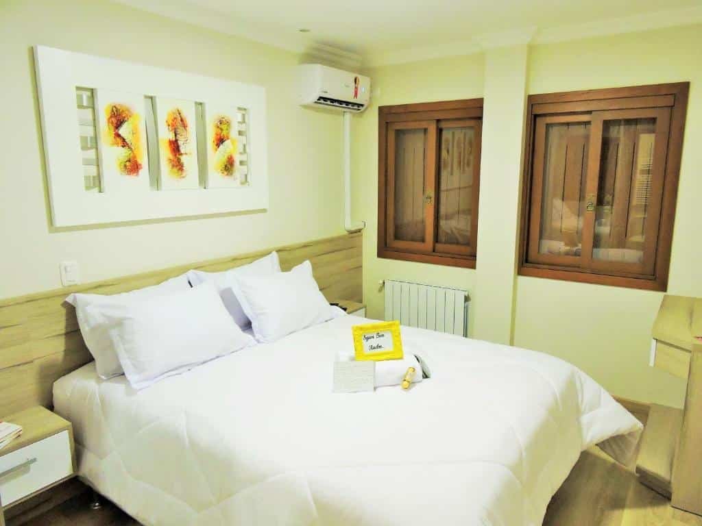Quarto em Apto no centro de Gramado, com duas janelas, um ar-condicionado, uma cama de casal com toalhas e travesseiros, um quadro e duas mesinhas, para representar airbnb em Gramado