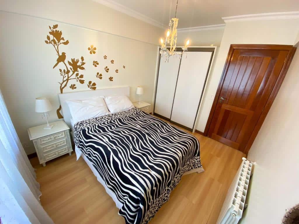 Quarto no Apartamento Primavera, a 600 metros da Rua Coberta, com 2 quartos, uma cama de casal, duas mesinhas com abajures, um armário e uma janela com cortinas, além de um lustre, para representar airbnb em Gramado