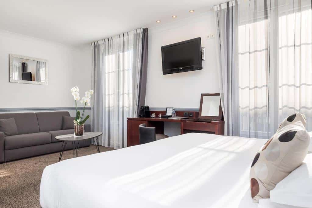 Quarto do Best Western Hotel Lakmi Nice. A cama está no lado direito do quarto, ao lado há uma parede com duas janelas grandes com cortinas e no meio uma mesa com cadeira e uma TV a cima. Em frente a cama há uma mesa de centro com uma flor e um sofá logo atrás. Imagem para ilustrar o post hotéis em Nice.