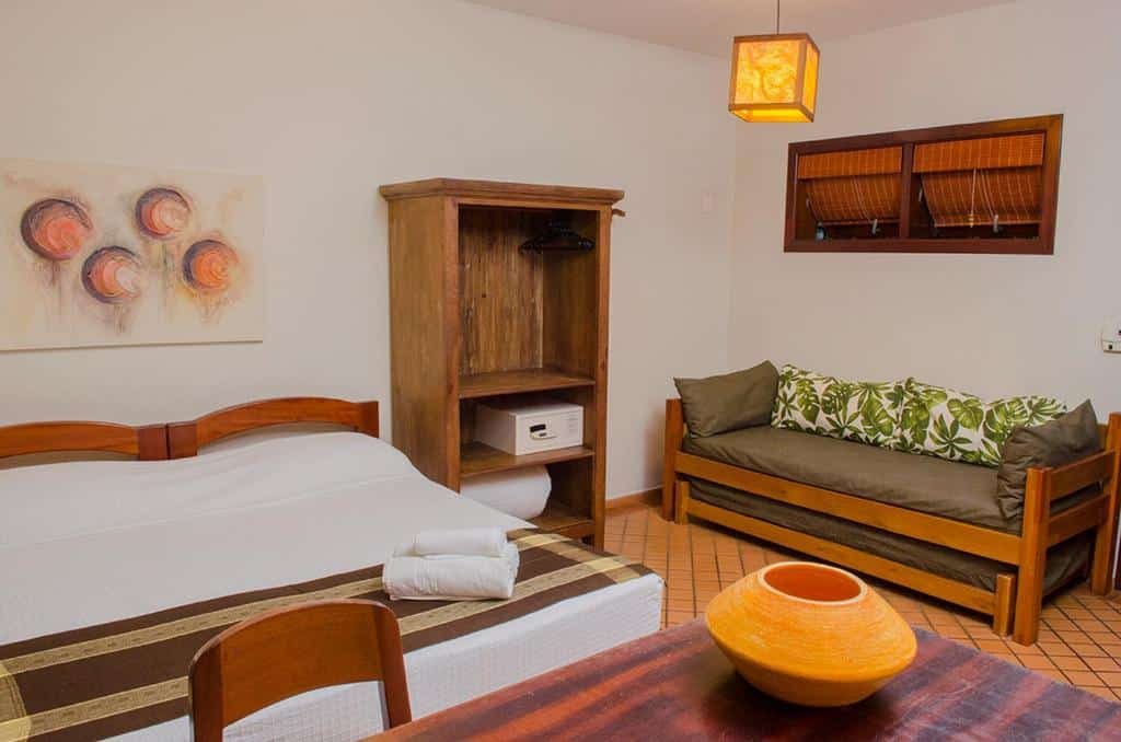 Quarto da Pousada Bico Verde com cama de casal ao centro, cômoda de madeira com cofre, sofá cama do lado direito e, a frente da cama, mesa de trabalho com cadeira.