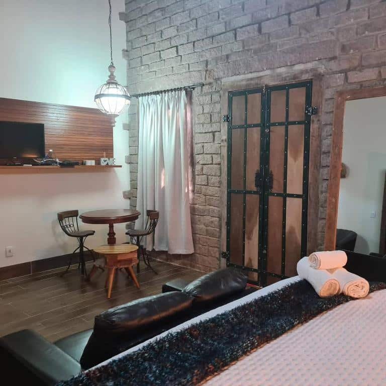suíte do Bosque São Francisco com uma parede de pedra, uma cama de casal, uma mesa com duas cadeiras e um painel de madeira com uma tv de tela plana