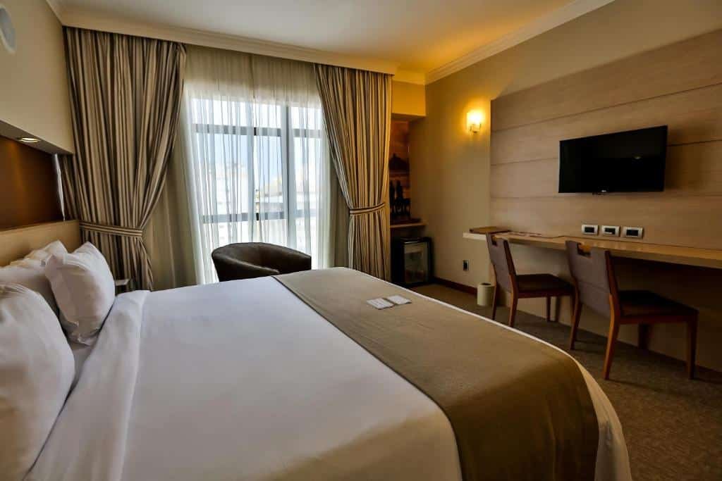 Quarto do Bourbon Santos Convention Hotel com uma varanda com cortinas, uma poltrona, uma cama de casal, uma bancada com duas cadeiras, além de uma televisão e um frigobar
