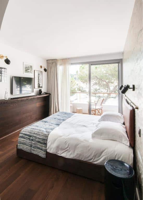 Quarto do Boutique Hotel Miramar. Uma cama de casal no lado direito, de frente para a cama quadros, luminárias e uma televisão. No fundo, a sacada do quarto.