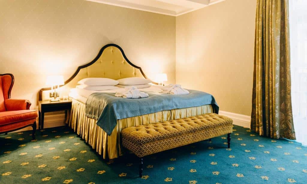 Quarto do Hotel Bristol com cama de casal com duas cômodas de cada lado da cama com luminárias e uma poltrona laranja do lado direito.
