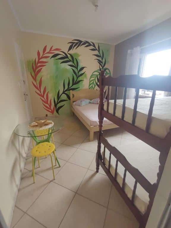 Quarto com cama de casal encostada em parede decorada do fundo do quarto, cama beliche na parede da frente, janela com cortina no lado direito e mesa de vidro com dois bancos na lado esquerdo. Imagem para ilustrar o post pousadas em Mongaguá.