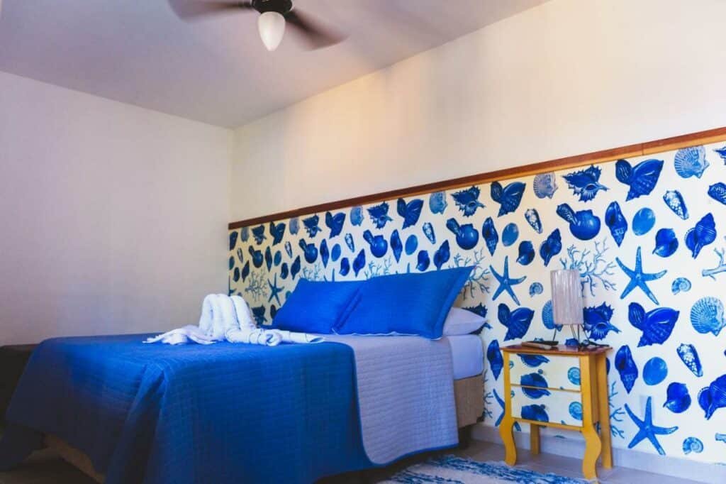 Quarto no Cantinho do PE com uma parede cheia de desenhos em azul de estrelas do mar e conchas, uma cama de casal, com toalhas e travesseiros sob ela, e uma pequena mesinha de cabeceira com um abajur