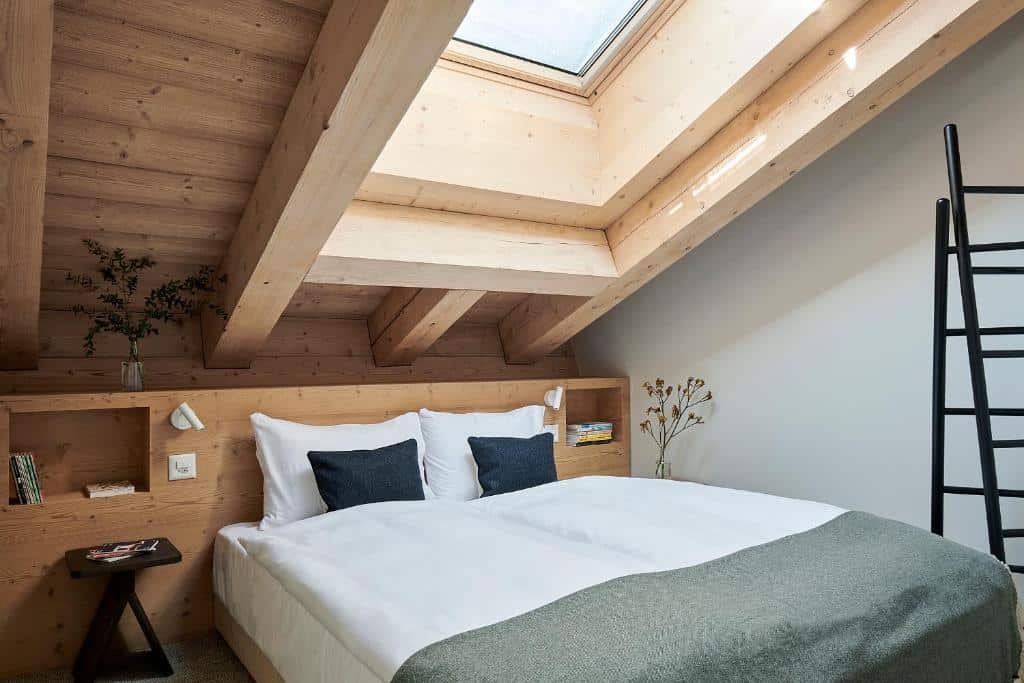 Quarto do Carina – Design&Lifestyle hotel com cama de casal do lado esquerdo da imagem com uma pequena cômoda em cada lado da cama e do lado esquerdo uma escada. Representa onde ficar em Zermatt.