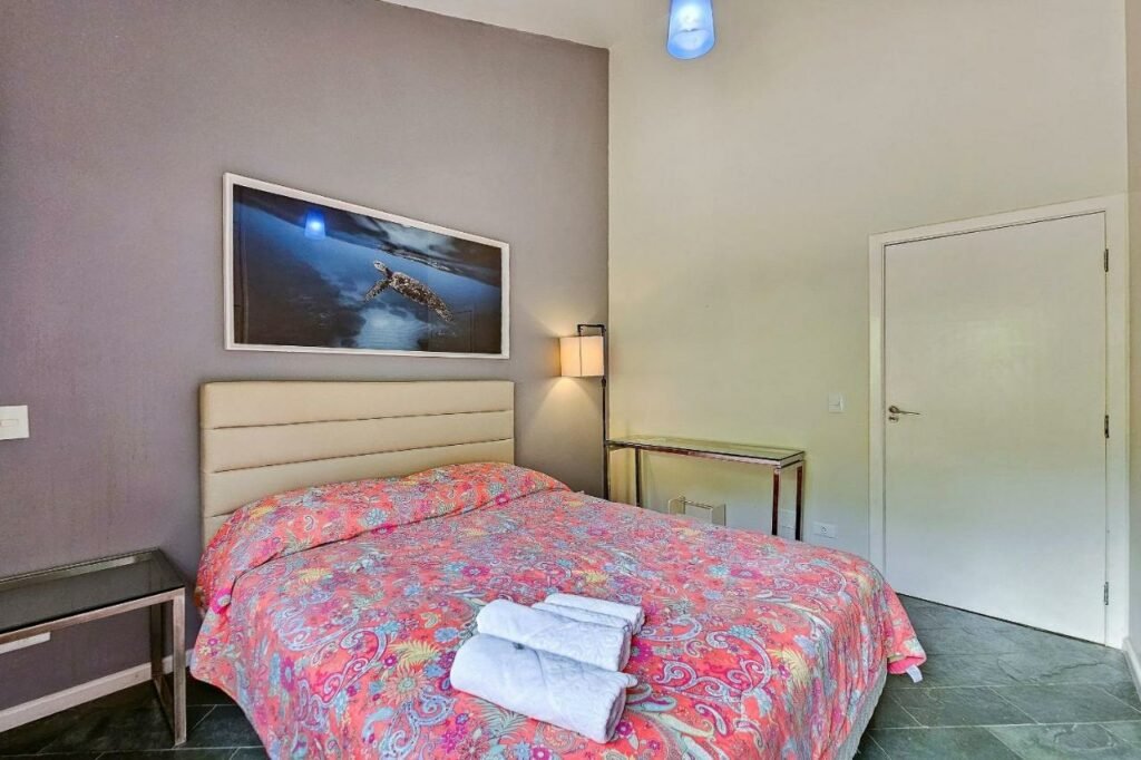 Quarto da Casa com varanda e churrasqueira em São Sebastião com cama de casal do lado esquerdo da imagem com uma mesa de vidro do lado direito da cama e do lado esquerdo da cama uma luminária e uma mesa de vidro. Representa airbnb na praia da Juréia.