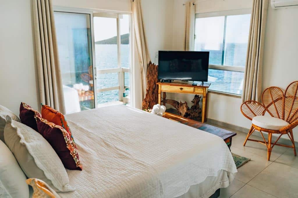 Quarto do airbnb Casa Grega. A cama de casal está encostada na parede do lado esquerdo da imagem e na frente da cama há um pequeno móvel com uma TV em cima e no lado direito há uma cadeira. No lado esquerdo do quarto há uma porta de vidro que dá acesso a varanda e possui vista para o mar. Imagem utilizada para ilustrar o post airbnb em Arraial do Cabo.
