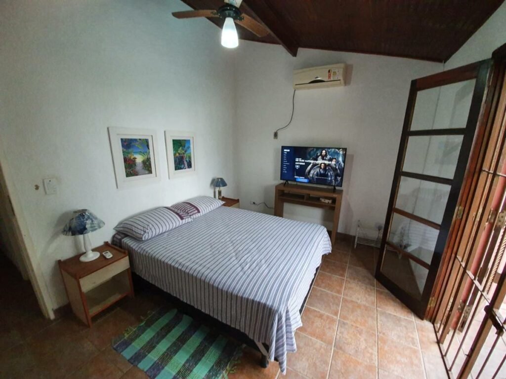 Quarto da Casa pé na areia em Toque Toque com cama de casal do lado esquerdo da imagem com uma cômoda em cada lado da cama com luminária do lado esquerdo da cama uma cômoda com TV. Representa airbnb na praia de Calhetas.