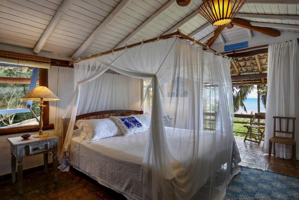 Quarto do Casas da Lia com cama de casal do lado esquerdo, com uma cômoda de madeira do lado esquerdo da cama com luminária e do lado esquerdo do quarto portas amplas de madeira com cortinas brancas que dá acesso à varanda.