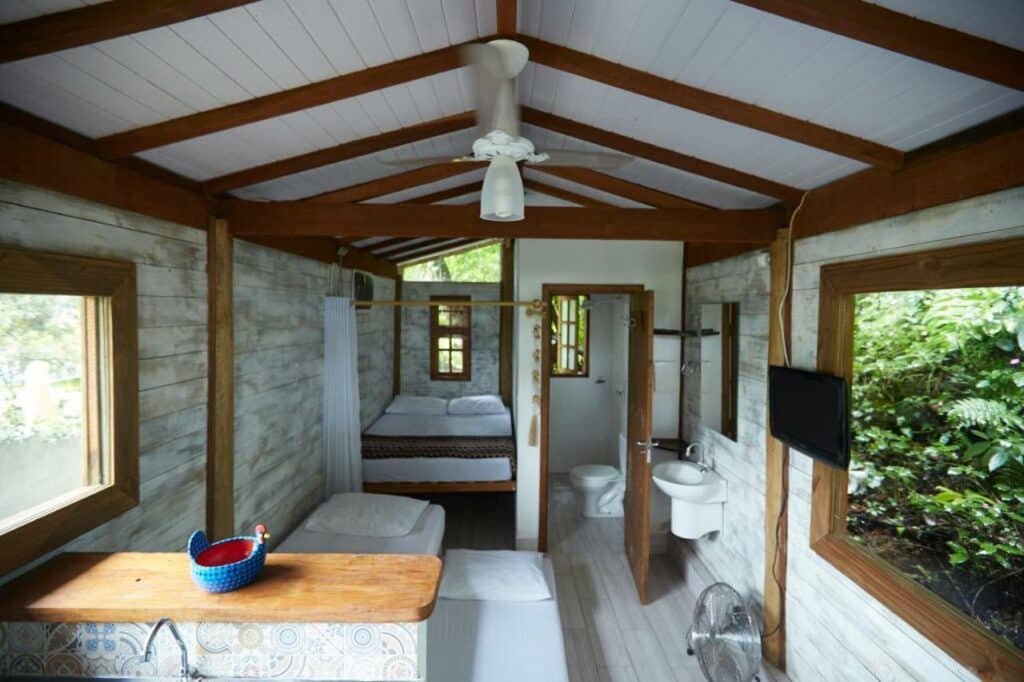 Imagem do quarto do Chalé Paúba 80m do Mar com um sofá cama do lado esquerdo da imagem e um colchão no chão e em frente uma TV pendurada na parede. Ao fundo uma cama de casal no lado esquerdo e do lado direito acesso ao banheiro.