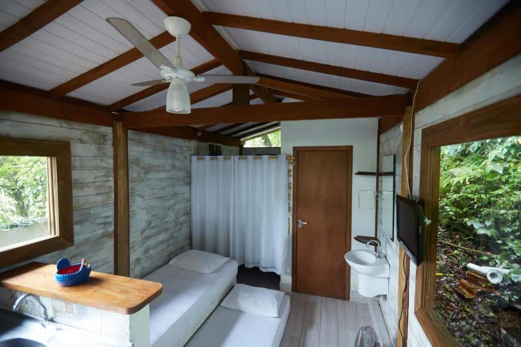 Vista do quarto do Chalé Paúba 80m do Mar com estilo rustico e simples com sofá cama na área central do quarto com um colchão no chão, pia do lado direito e acima TV na parede e ao lado janela de vidro.