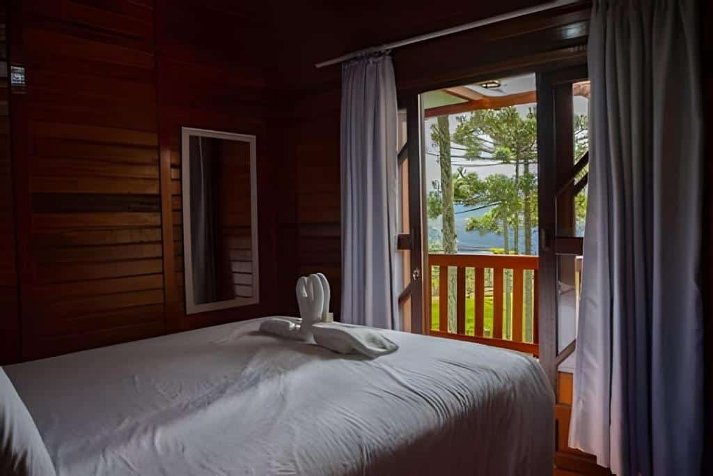 Quarto do Chalé Urubici - Morada nas Montanhas com cama de casal, cisnes feitos de toalha em cima da cama se beijando, espelho de corpo inteiro e varanda de madeira com vista de natureza.
