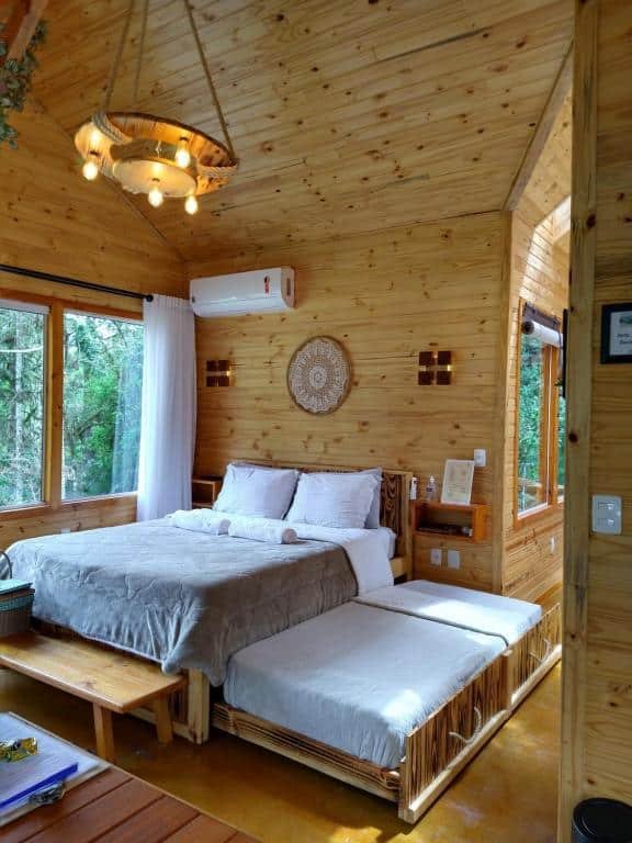 Vista do quarto do Chalés Estância Cachoeira do Avencal vom cama de casal e uma cama extra ao lado no chão, há um ar-condicionado e ma janela bastante ampla de vidro com vista da natureza ao lado com decoração rústica.