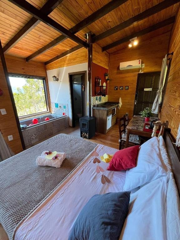 vista do interior do quarto dos Chalés incríveis com banheira de hidromassagem e vista encantadora com cama grande no centro com banheira à frente, com janela acima, do lado, há uma mesa com cadeiras de madeira e uma pequena cozinha com frigobar e micro-ondas, sendo uma das opções de chalés em Urubici.
